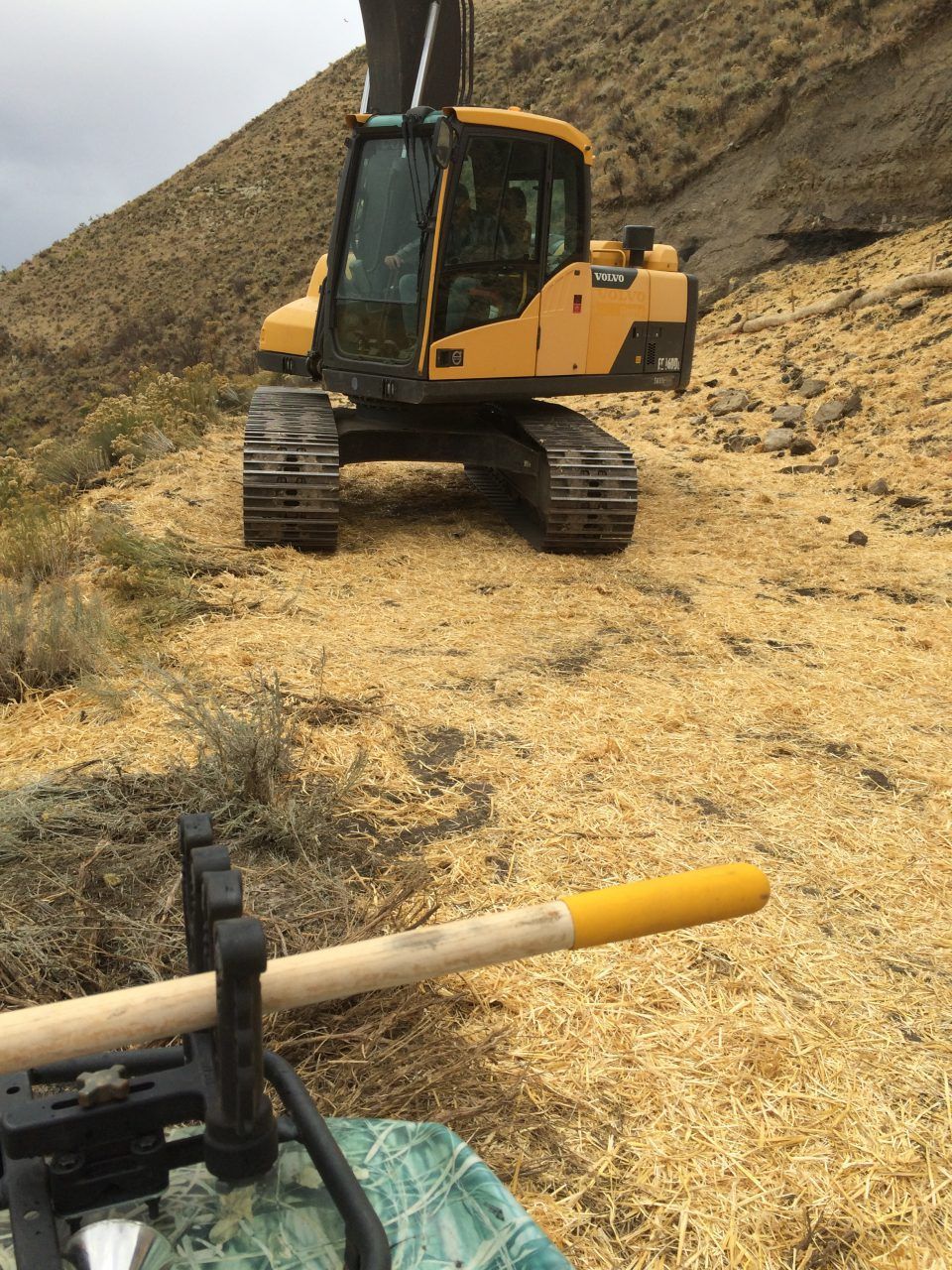 Yellow excavator on a hay-covered slope, with a farm tool visible in the foreground.