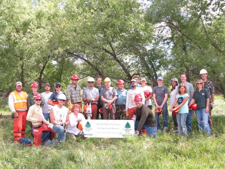 Group of volunteers in helmets and safety gear posing with a banner in a wooded outdoor area