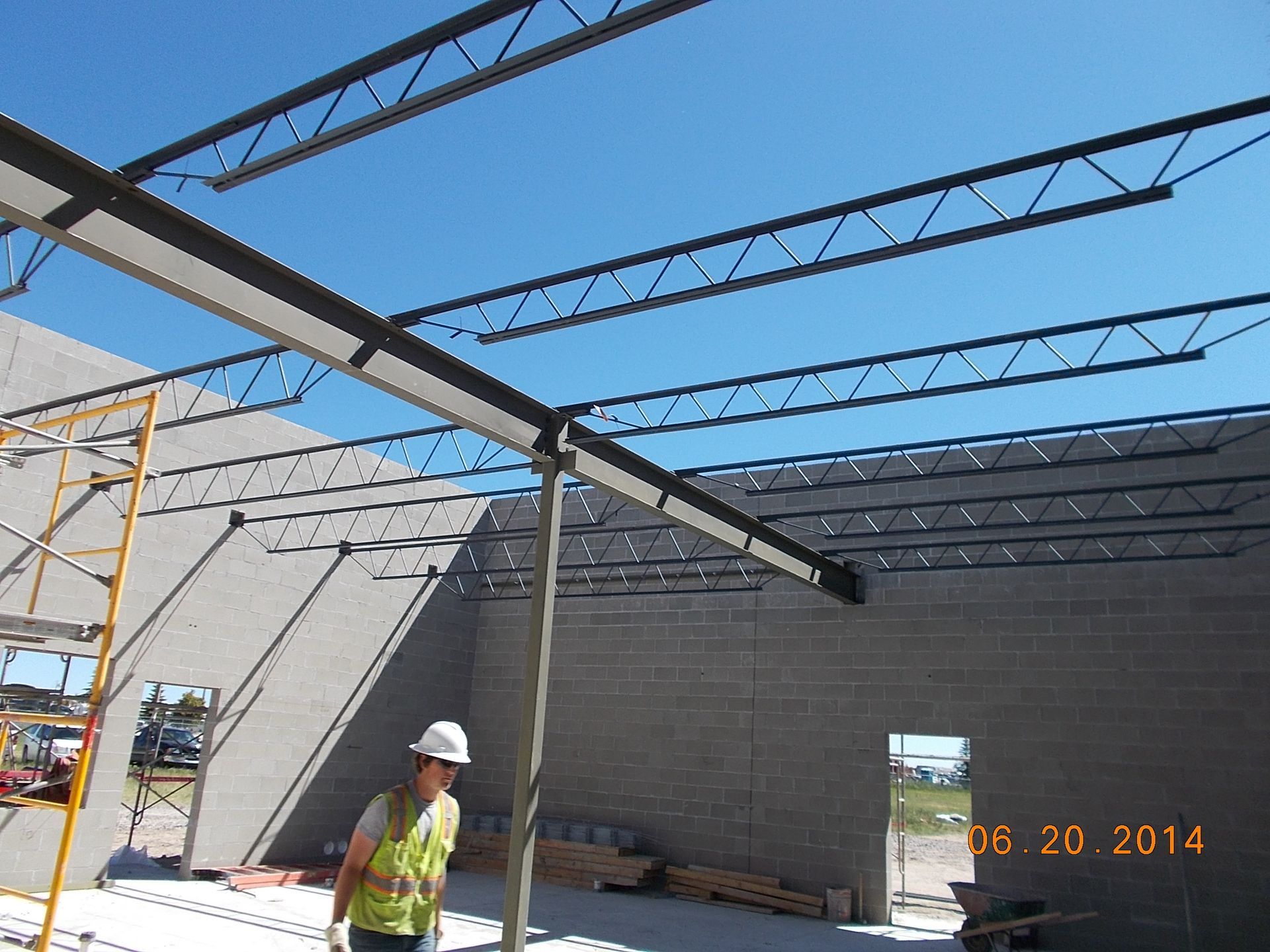 Construction worker under steel roof framing at a building site, with gray block walls and blue sky.
