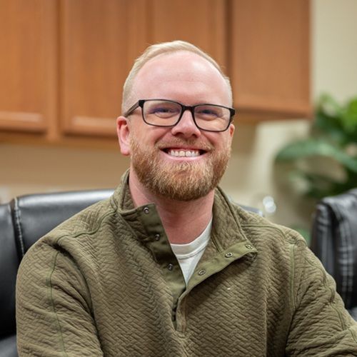 Smiling person in glasses and green sweater seated in an office chair in a wood-paneled office