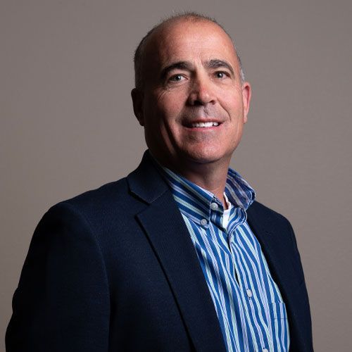 Studio portrait of a smiling man in a navy blazer and blue striped shirt against a gray background