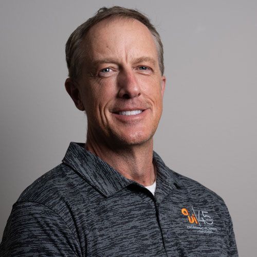 Portrait of a smiling man in a gray collared shirt with an orange logo on a plain gray background