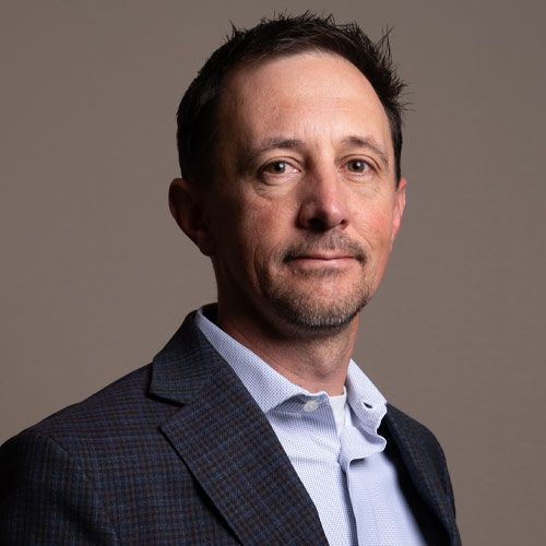 Studio headshot of a man in a dark blazer and light blue shirt, facing the camera against a gray background