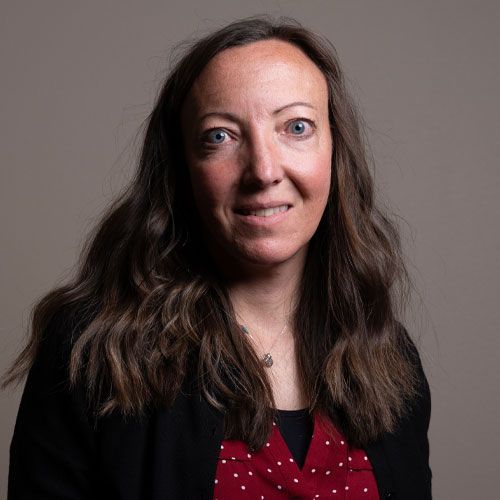 Portrait of a woman with long dark hair wearing a black jacket and red polka-dot blouse on a gray background