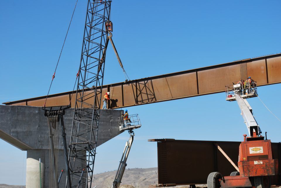 Workers using lifts to assemble a large steel bridge span at a construction site under a blue sky