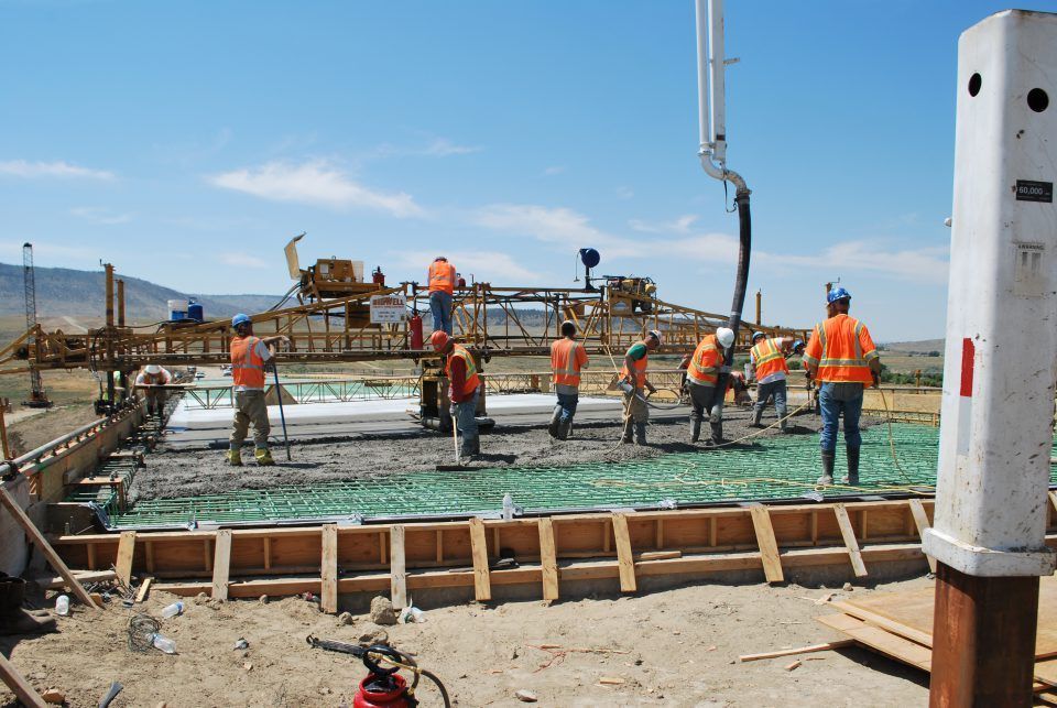 Construction workers in orange vests pour concrete at a building site under a clear blue sky.