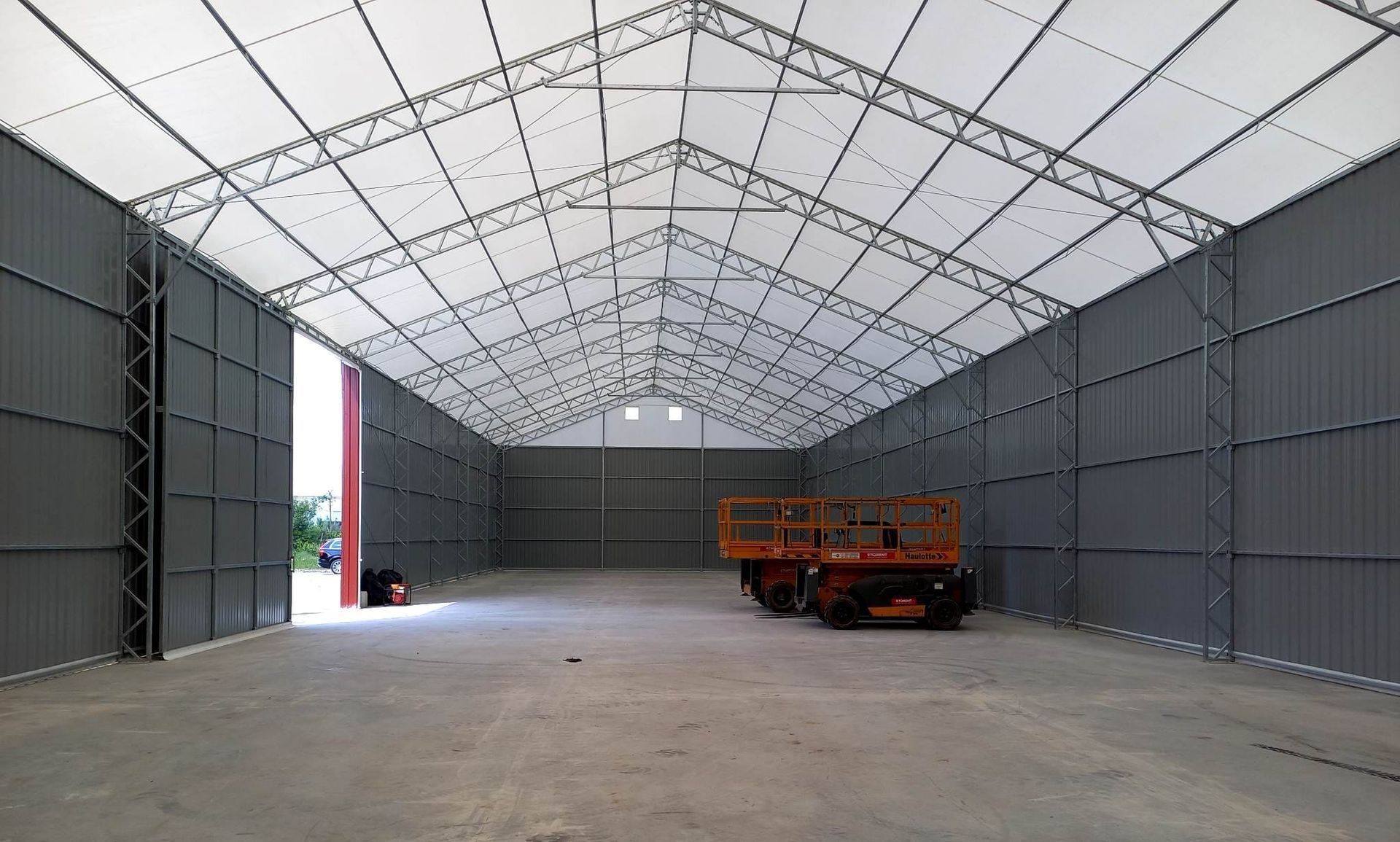 Tuščias pramoninis sandėlis su baltu stogu ir pilkomis sienomis; viduje yra raudona transporto priemonė.