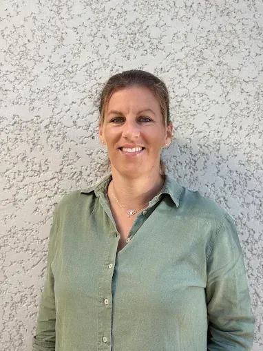 Femme souriante en chemise verte boutonnée, debout contre un mur à la texture claire