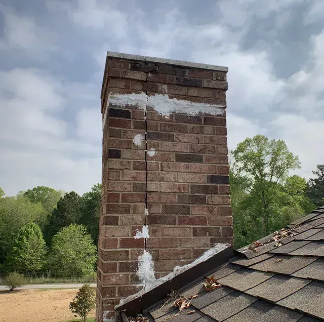 Brick chimney above a shingled roof under a cloudy sky, with trees in the background
