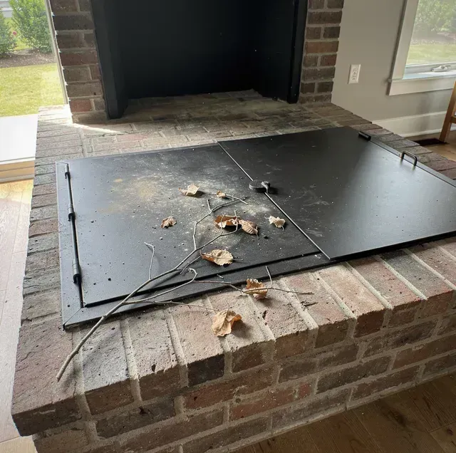 Black fireplace hearth with a metal cover, cords, and leaves on a brick surround.