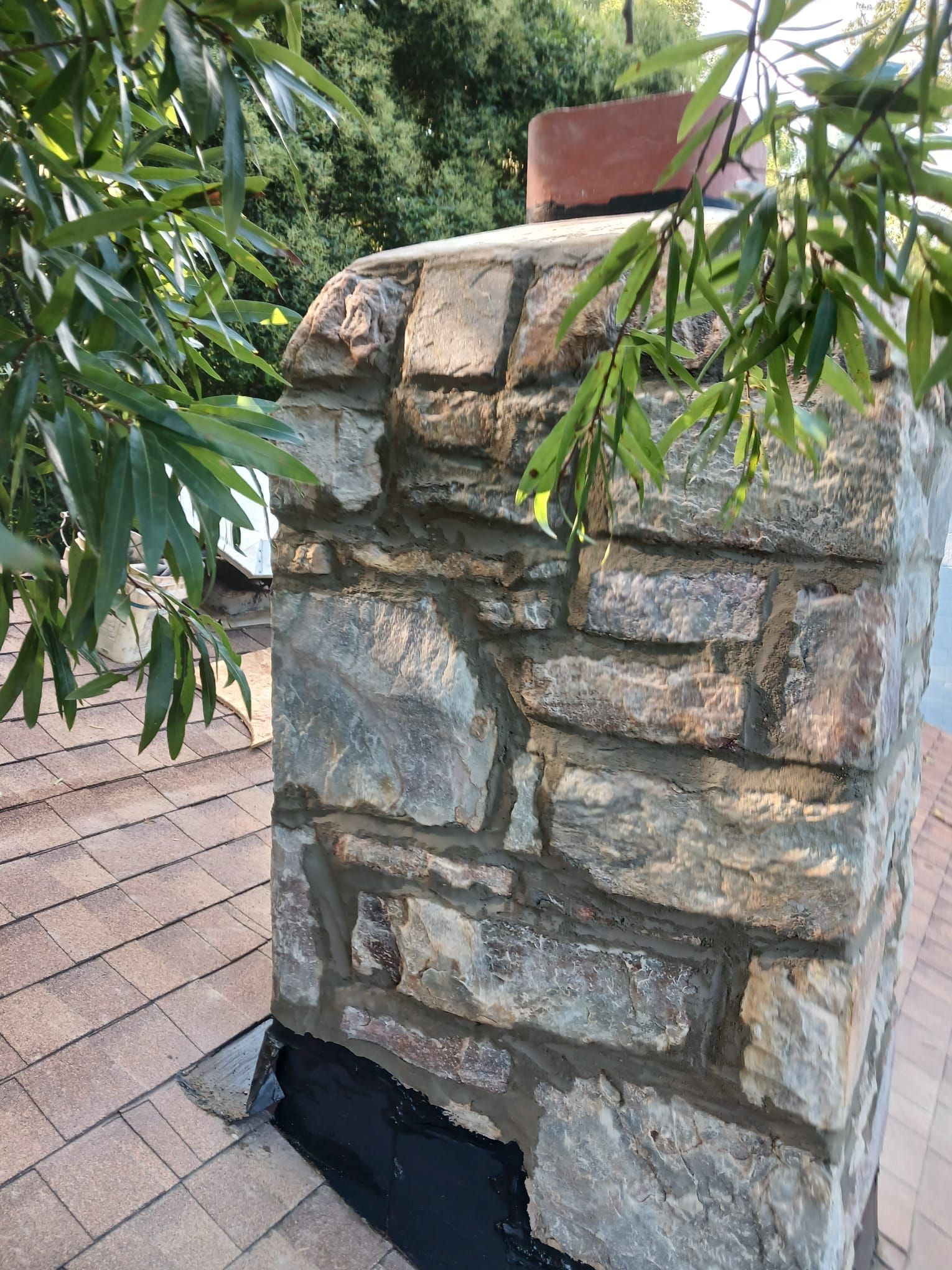 Stone chimney on a shingled roof with overhanging green leaves and a red cap on top