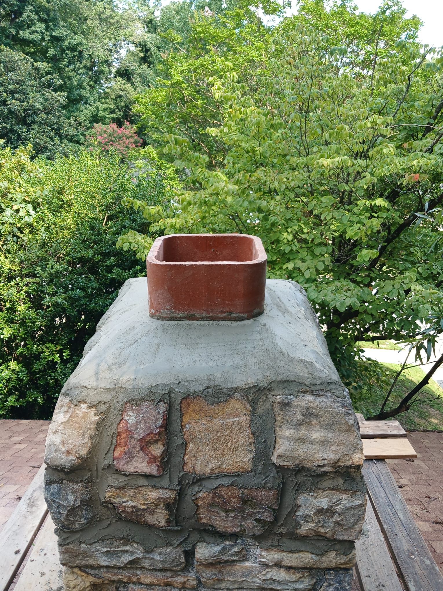 Brick chimney cap with a red clay flue pipe against leafy green trees