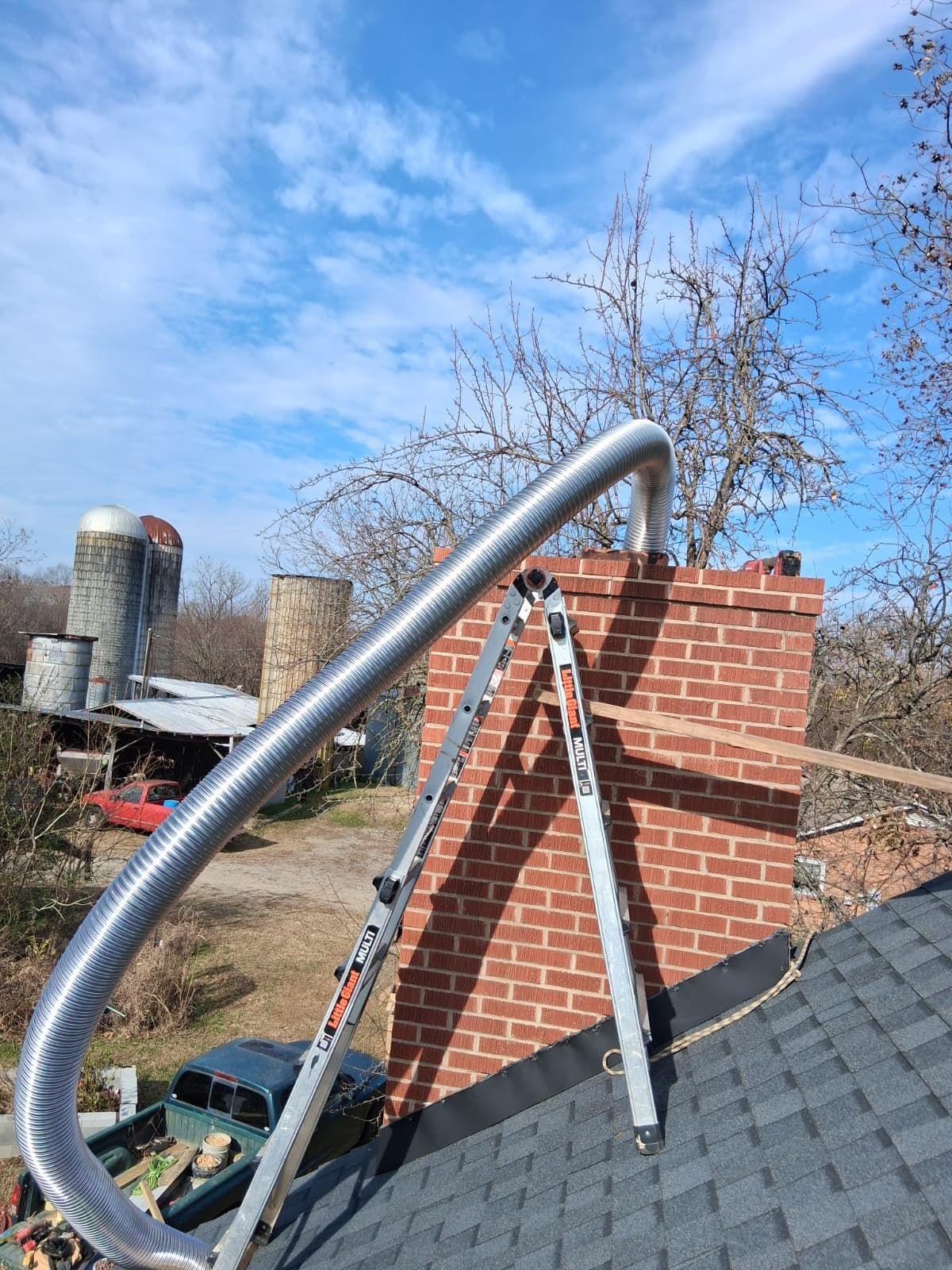Silver flexible duct extends over a red brick chimney on a rooftop under a blue sky.