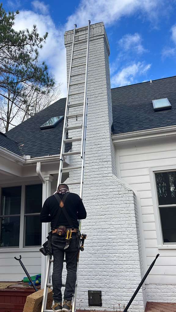 Worker on a ladder cleaning a tall white chimney beside a house with a dark roof.