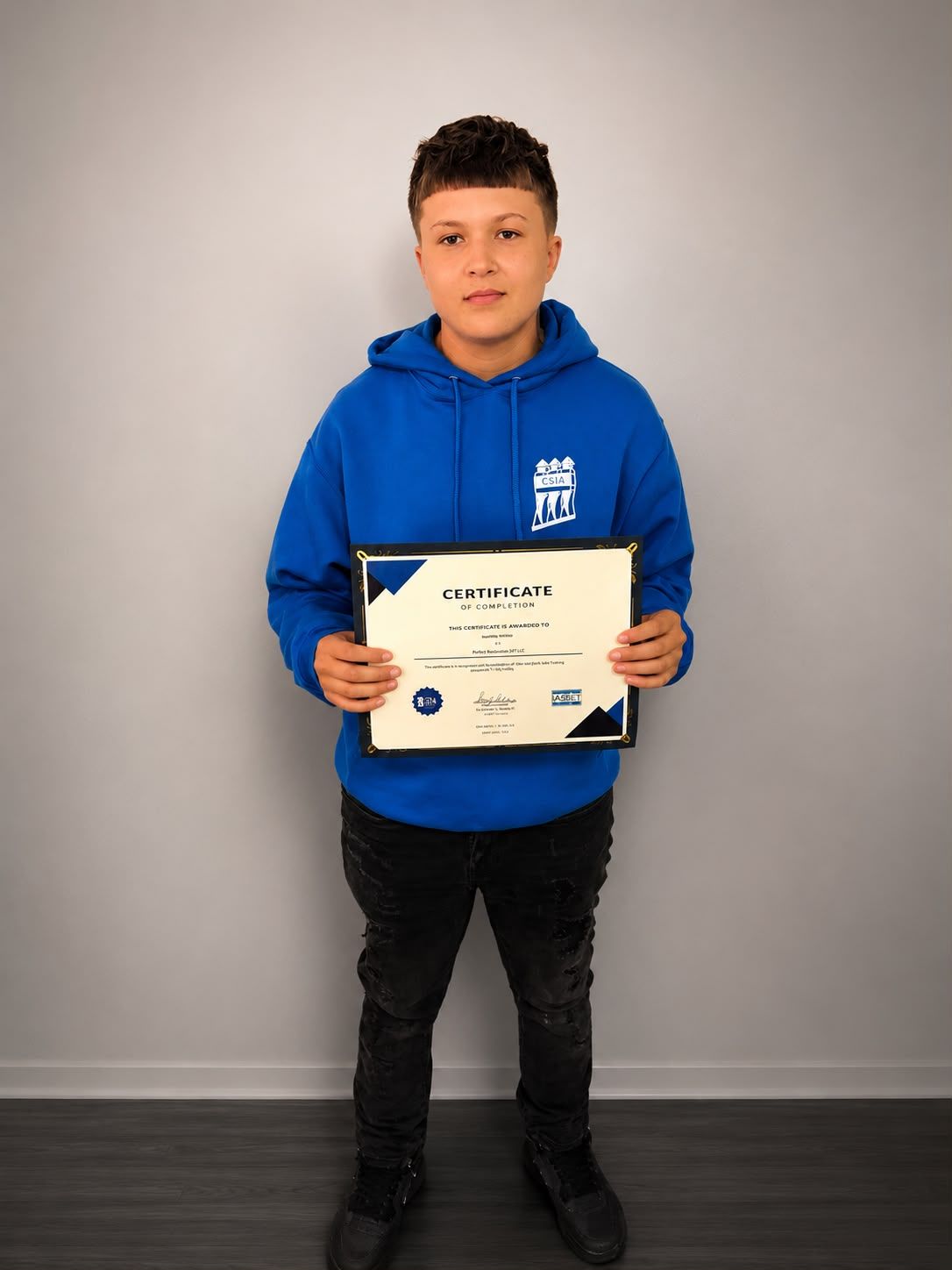 Person in a blue hoodie holding a certificate against a plain gray wall.