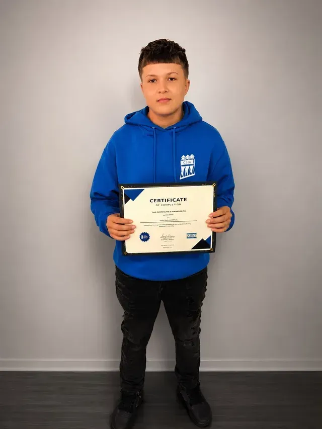 Person in a blue hoodie holding a certificate against a plain gray wall.