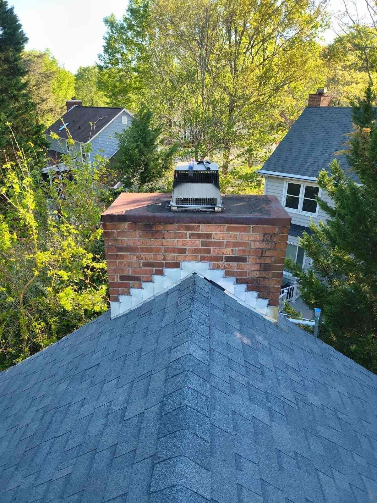 Rooftop view of a brick chimney and gray shingle roof surrounded by green trees and neighboring houses