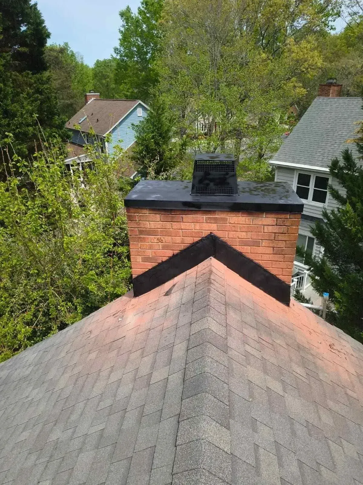 View over a shingled rooftop to a brick chimney, surrounded by leafy trees and neighboring houses