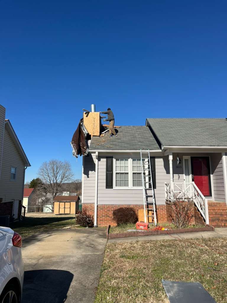 Damaged roof with exposed plywood on a suburban house beside a driveway under a clear blue sky