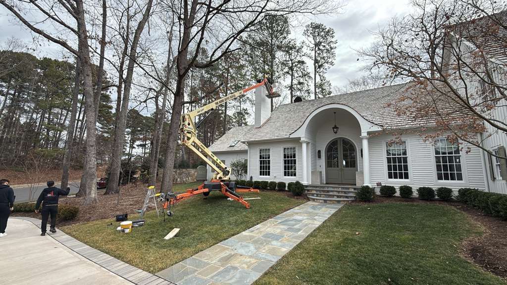 Construction crew using a boom lift to trim trees beside a white house with an arched porch.