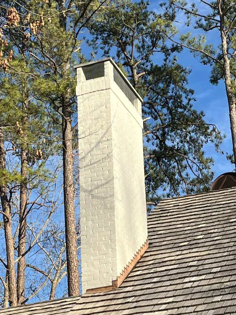 Tall white chimney on a shingled roof against blue sky and trees