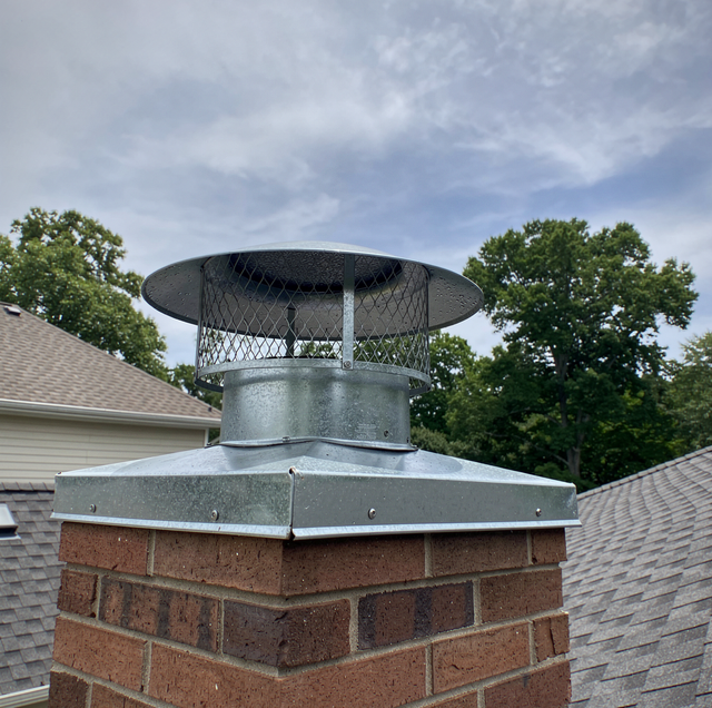 Metal chimney cap on a brick rooftop chimney against a cloudy sky and trees