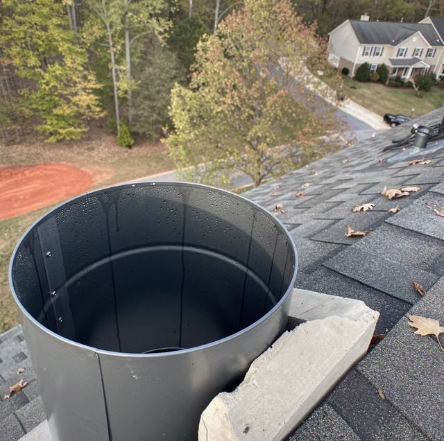 Black roof vent pipe beside shingles on a sloped roof, with trees and houses in the background.