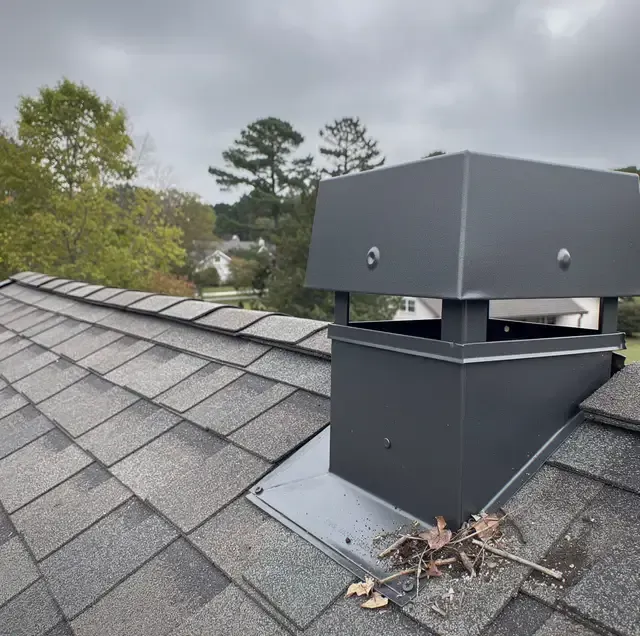 Dark gray rooftop vent box on a shingled roof with trees in the background