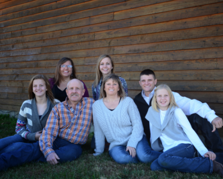 Seven people posing outdoors against a wooden wall, seated and standing on grass, smiling at the camera