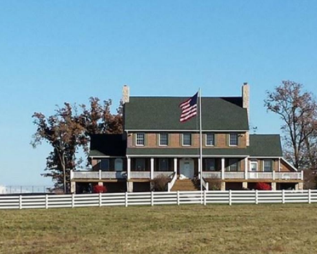 Two-story farmhouse with an American flag, front porch, and white fence on a grassy field under a clear sky