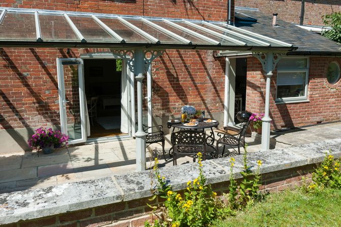 Patio outside a brick house with a glass awning, table and chairs, and flowers by the doorway