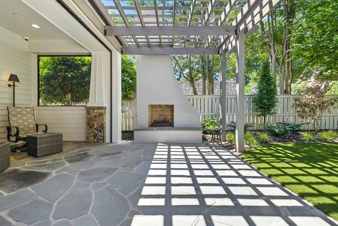 Covered patio with stone floor, pergola shadows, and a fireplace opening beside green trees and a fence