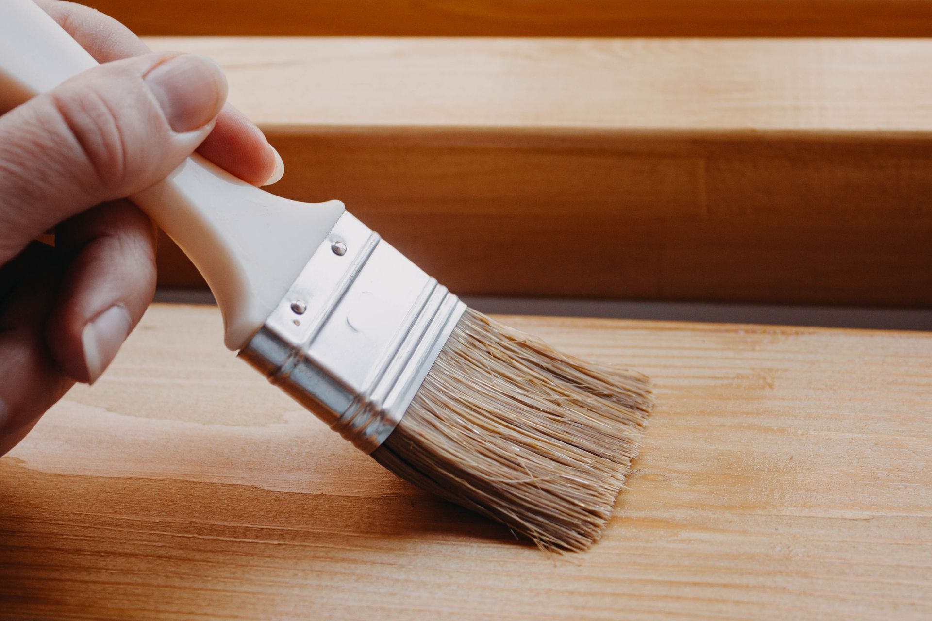 Mano che impugna un pennello mentre applica una finitura trasparente su una superficie di legno.