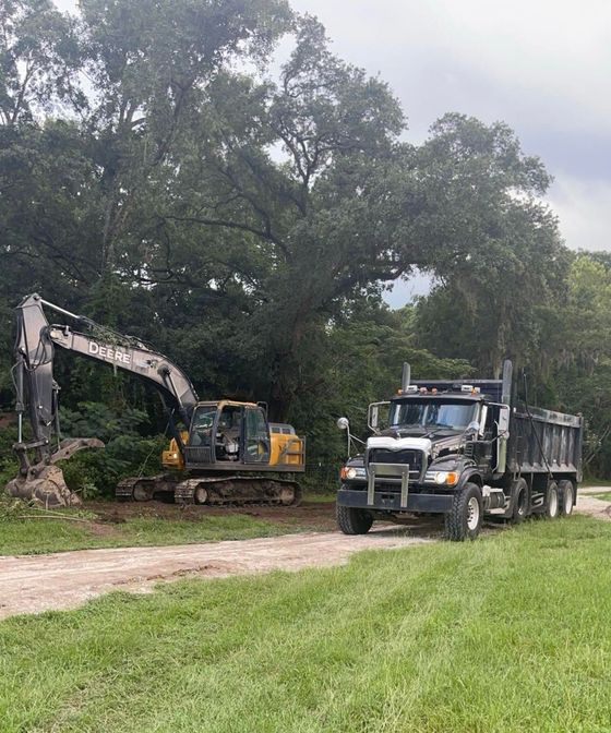 Excavator and dump truck at a dirt road work site beside large trees