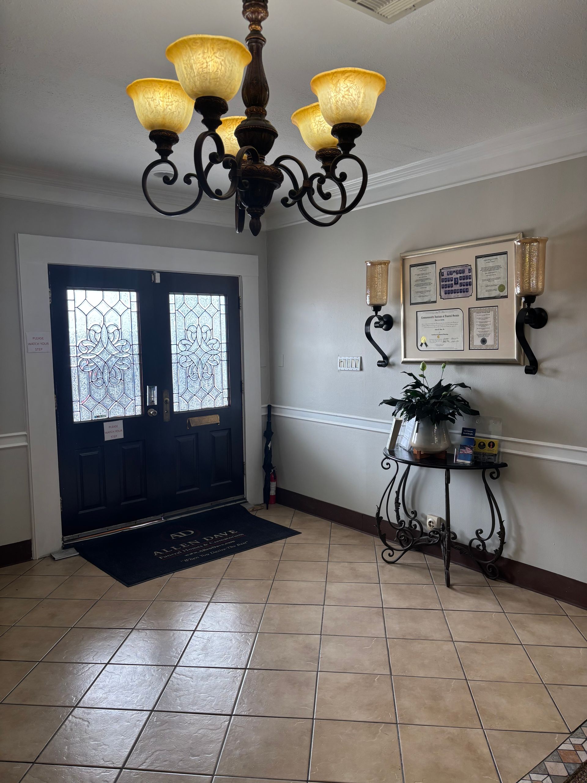 Elegant foyer with black double doors, chandelier, tiled floor, and a side table with plants and wall art