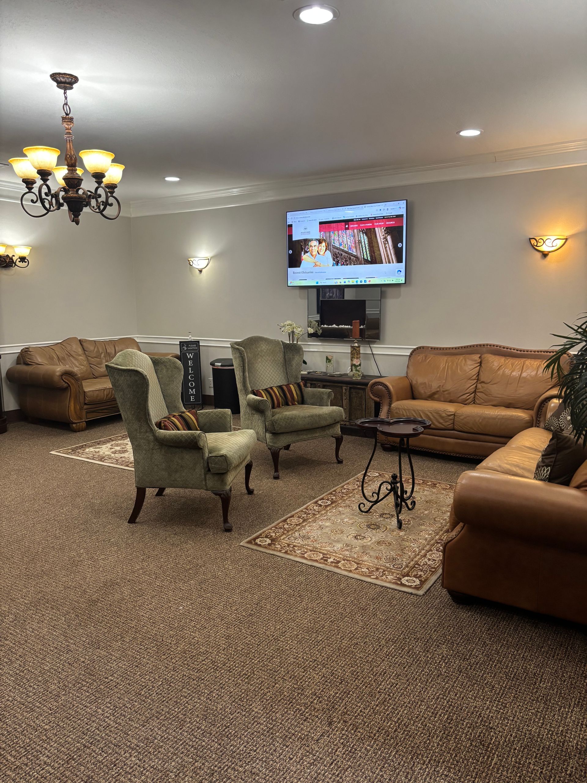 Cozy living room with tan sofas, armchairs, chandelier, and a wall-mounted TV above a fireplace.