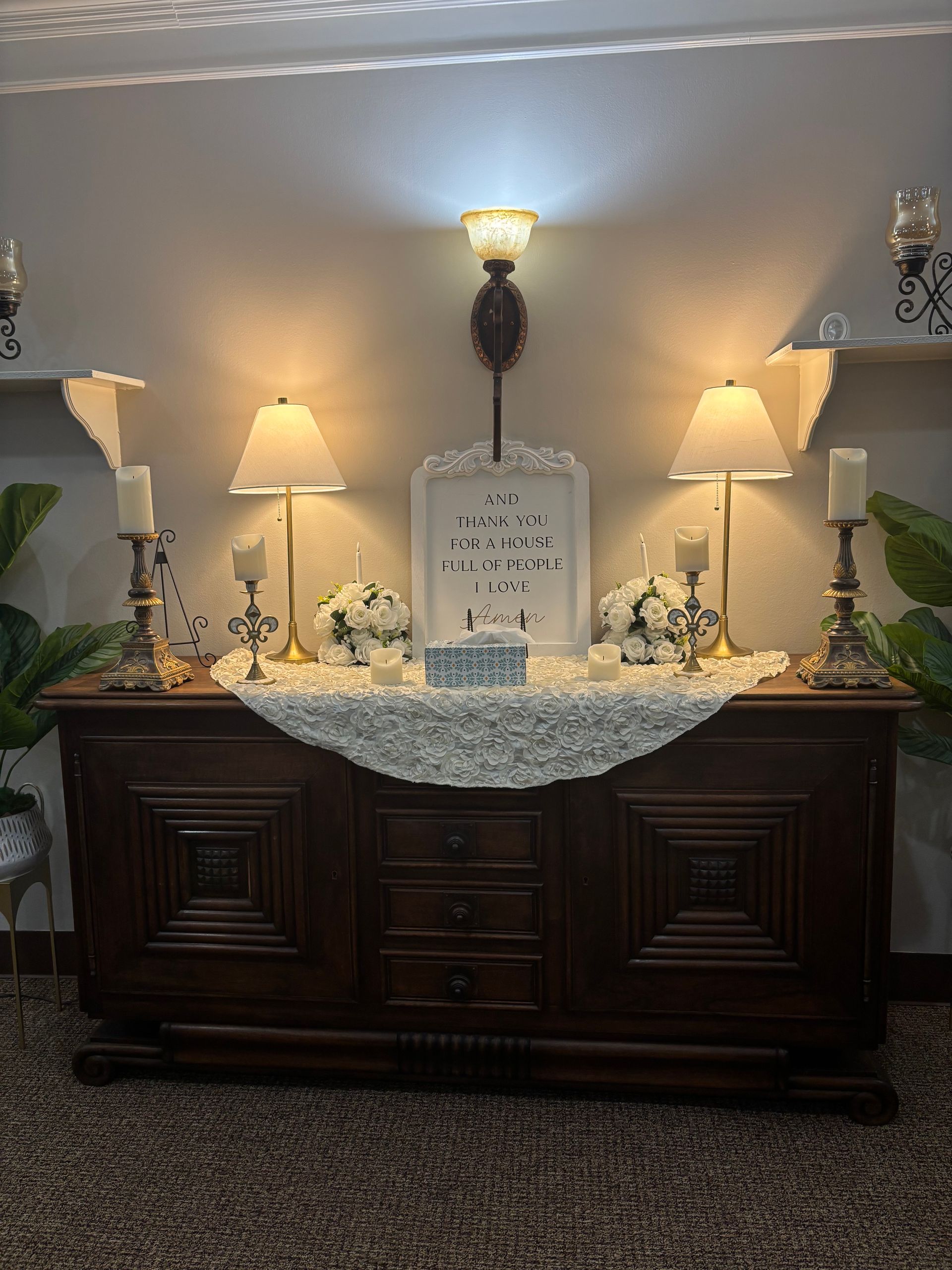 Decorated memorial table with candles, flowers, and a framed sign under wall sconces