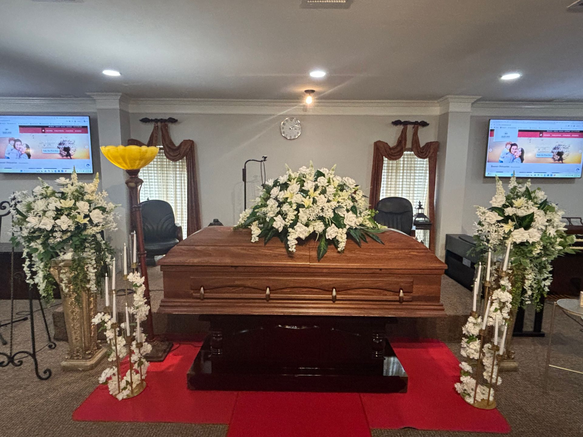Flower-adorned wooden casket in a funeral chapel with red carpet and framed screens on either side