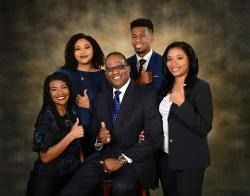 A group of five people in professional attire posing against a dark, textured backdrop, all giving a thumbs-up gesture.