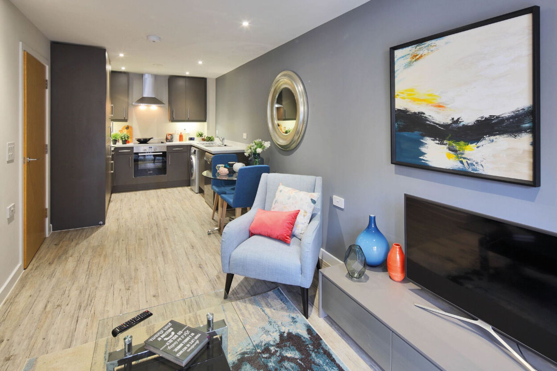 Modern living room with gray walls, blue armchair, TV, wall art, and open kitchen in the background