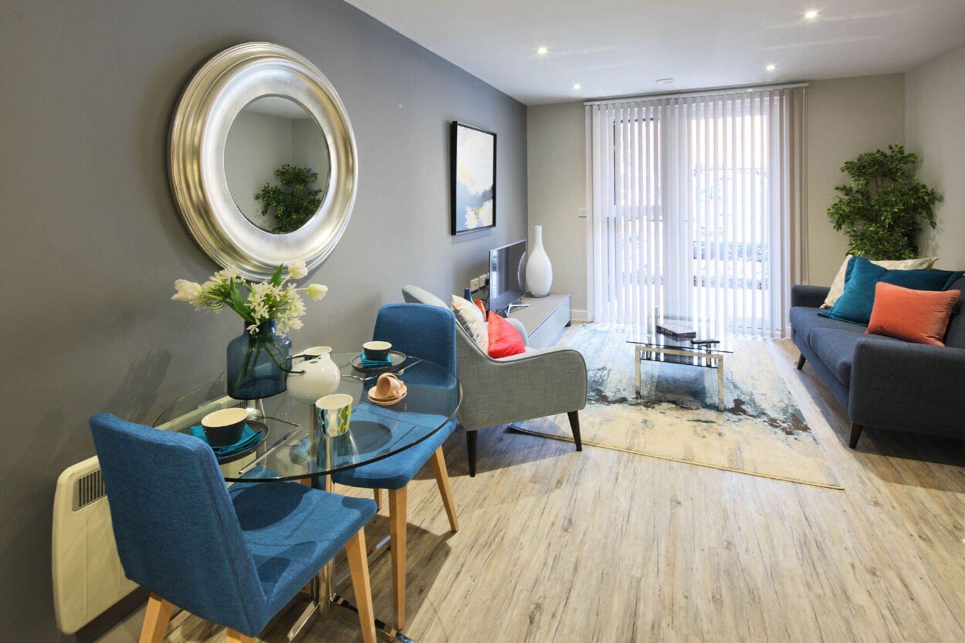 Bright living room with blue chairs, gray sofa, glass table, and sliding doors to a balcony.