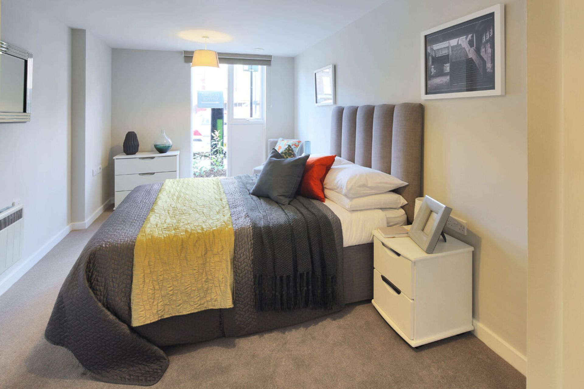 Bright bedroom with a bed, bedside tables, framed art, and a window at the far end.