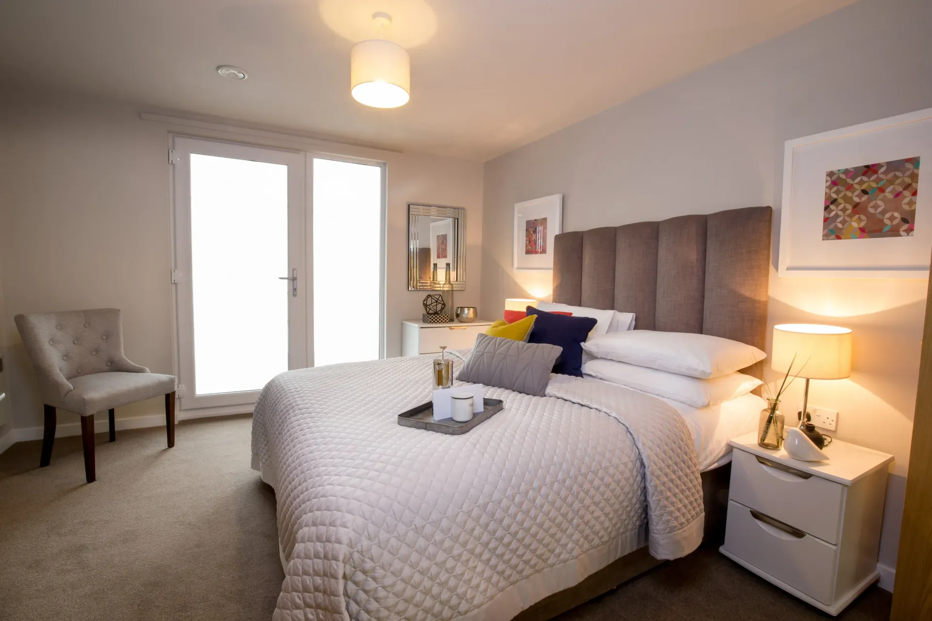 Bright bedroom with a made bed, bedside lamps, chair, and French doors to a balcony.