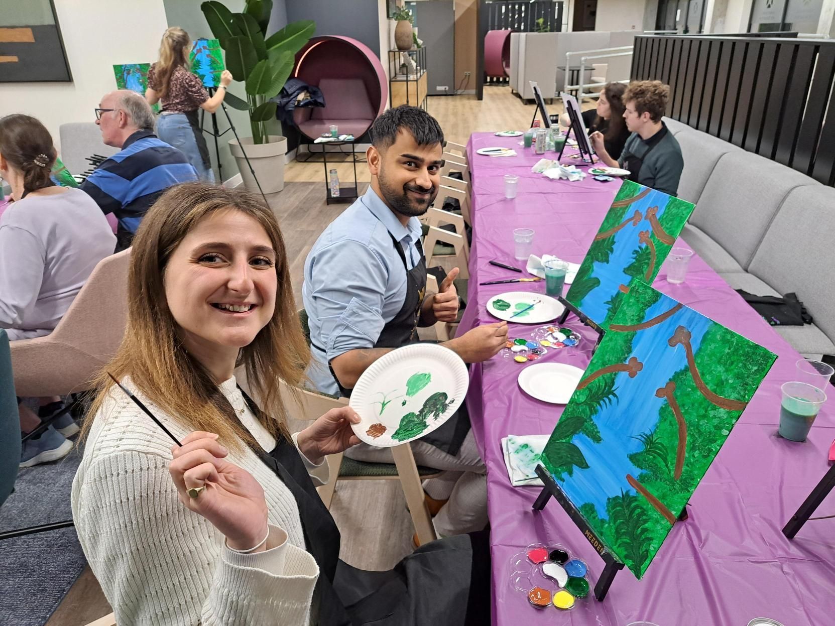 People painting at a table, smiling with palettes and colorful canvases in a community art setting