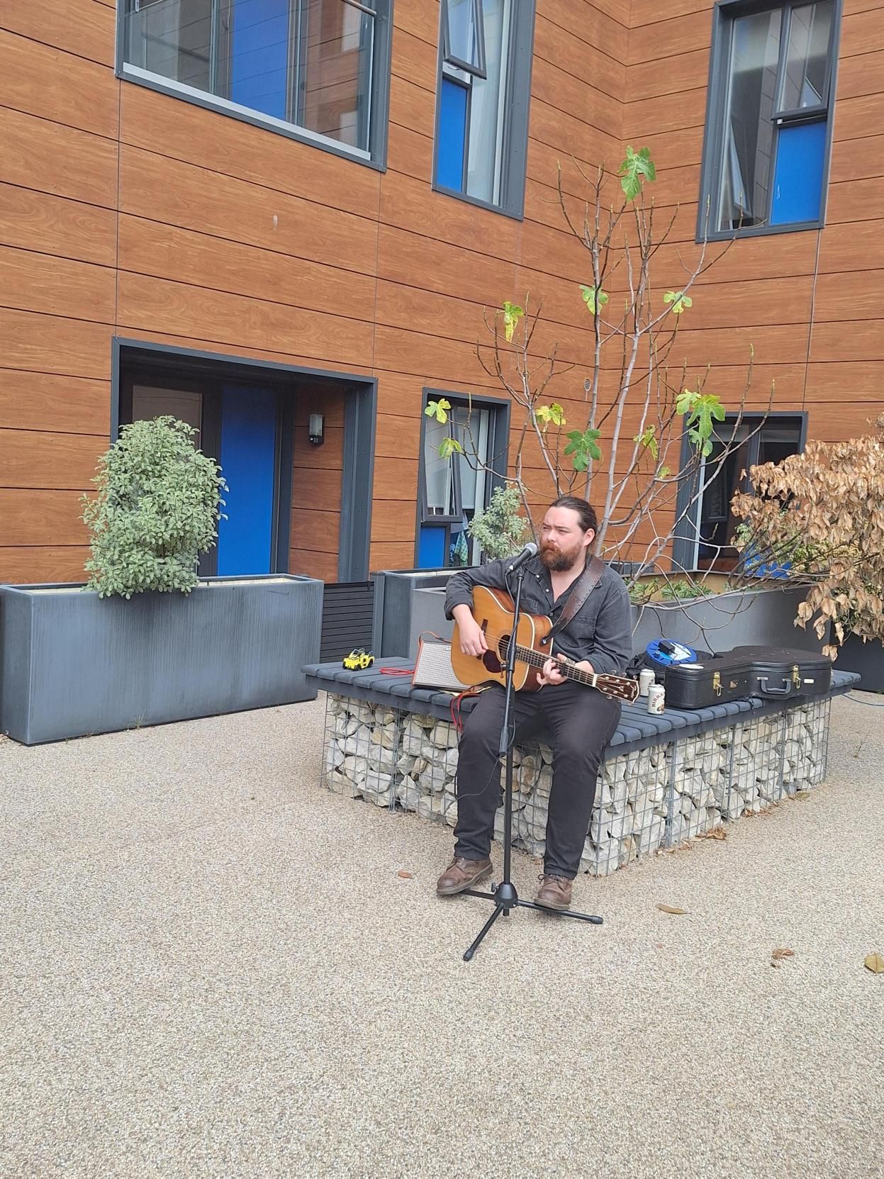 Man playing guitar on a courtyard patio beside a brick building with blue doors and windows