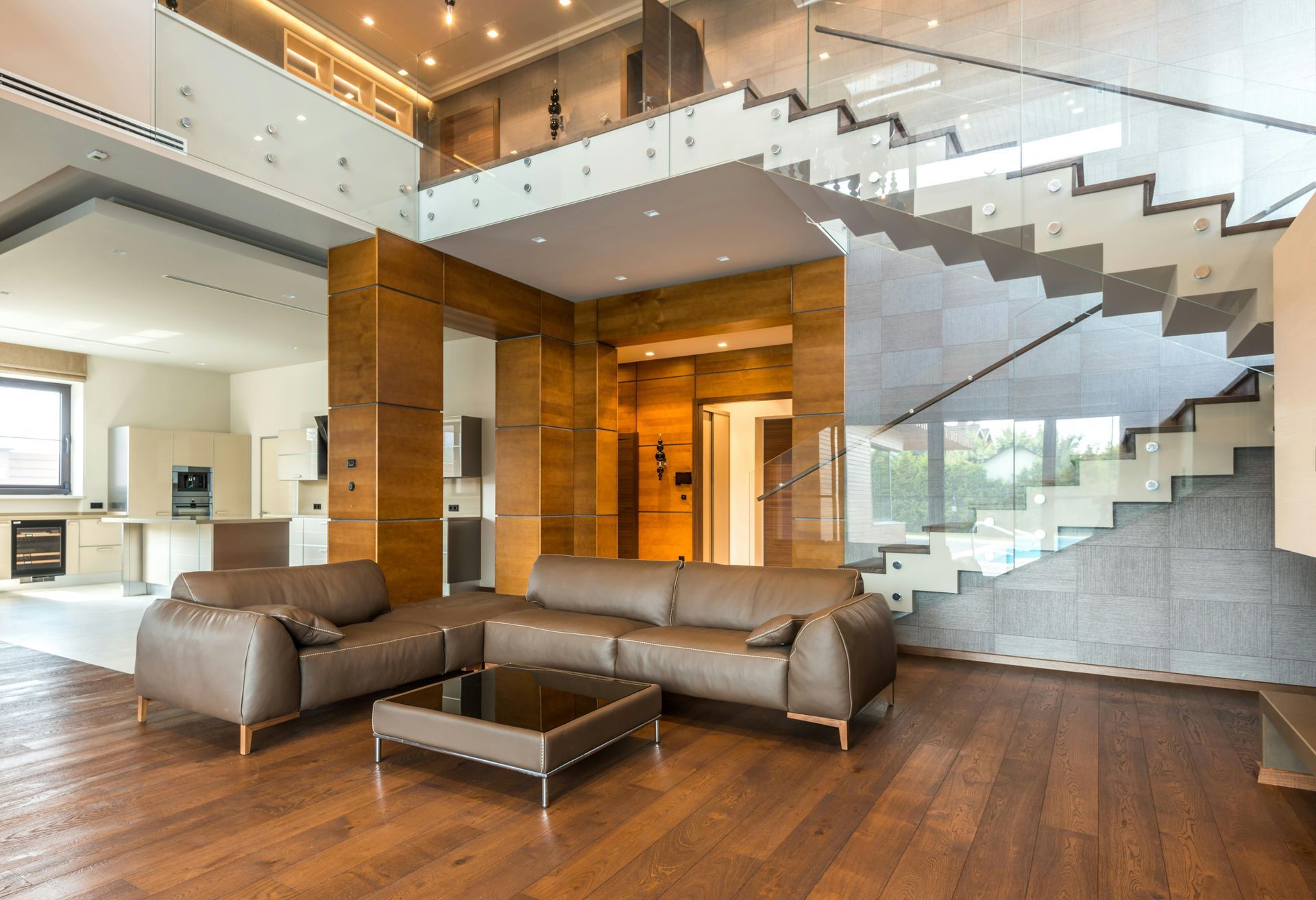 Modern living room with leather sectional, wood floors, glass staircase, and open kitchen.