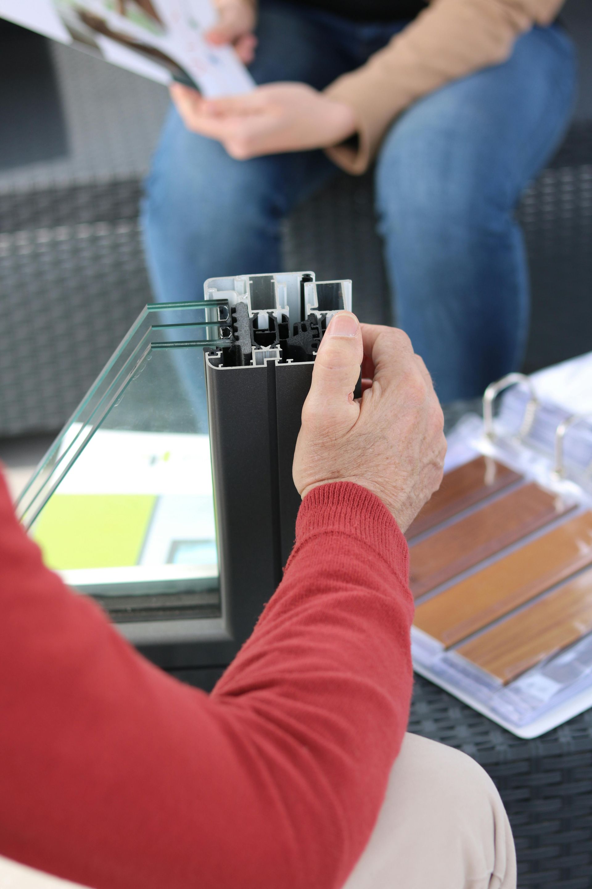 Person in red sweater examining a window cross-section, with another person and samples nearby.