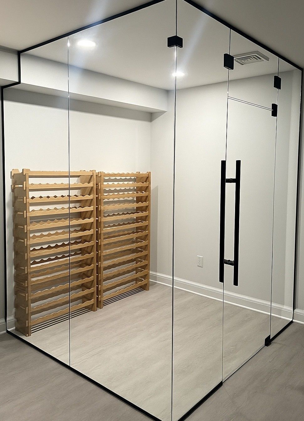 Glass-enclosed wine cellar with wooden racks against a pale wall, black-framed glass door, and recessed lighting. Birmingham, MI.