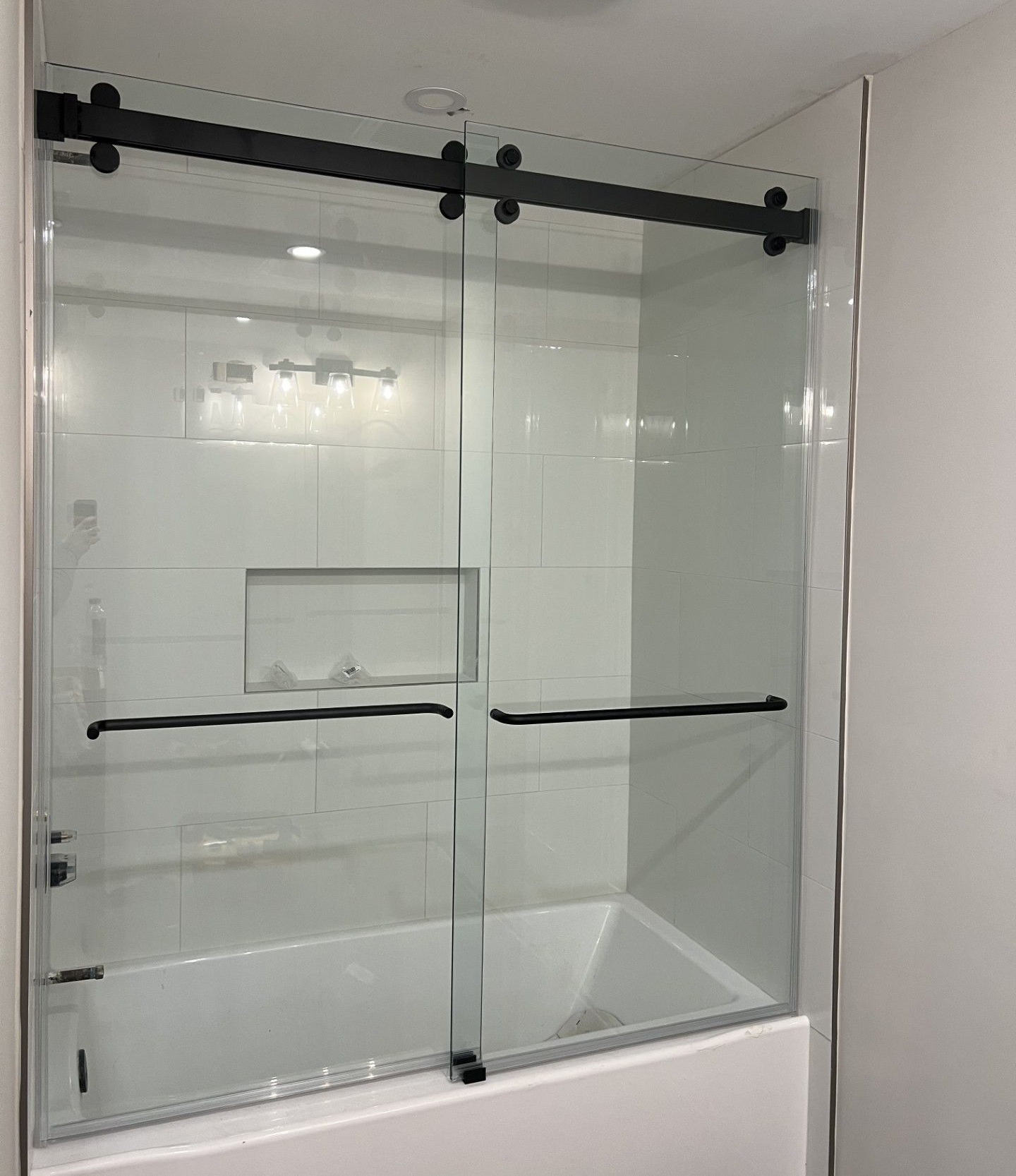 A glass shower door with black hardware installed over a white bathtub. The shower features a sliding door system.