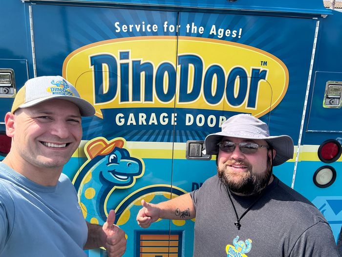 Two smiling men standing in front of a colorful DinoDoor garage door sign.
